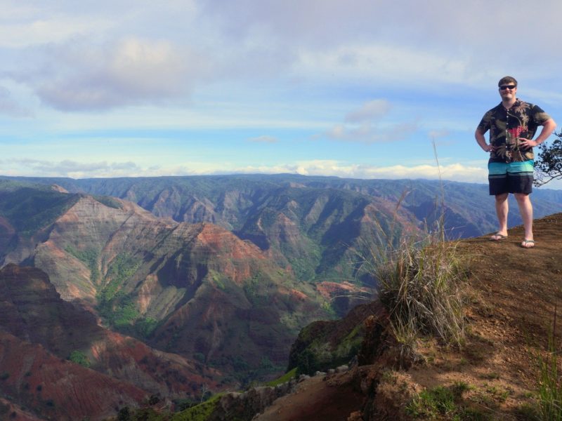 A person stands confidently at the edge of a cliff, overlooking the vibrant and colorful landscapes of Waimea Canyon with mountains and valleys stretching into the distance.