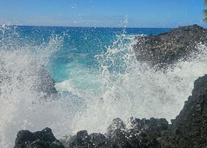 A striking view of ocean waves crashing against dark rocks, surrounded by clear blue water and a bright sky.