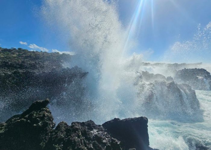 A powerful wave splashes against dark rocky formations under a bright blue sky, showcasing the dynamic interaction between water and land.
