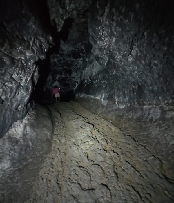 A person walks through a dark lava tube cave illuminated by a flashlight, showcasing rugged rock walls and uneven ground.