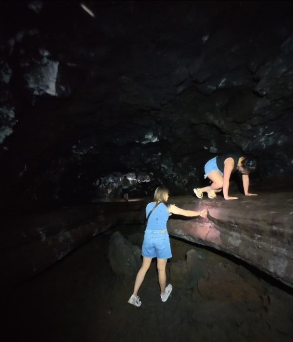 Two women navigate rocky terrain within a dimly lit lava tube cave while one uses a flashlight for visibility.