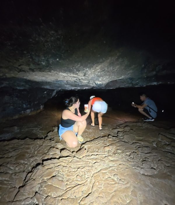 Three people exploring a dimly lit cave with rocky terrain, one crouching while another stands and the third takes a photo.