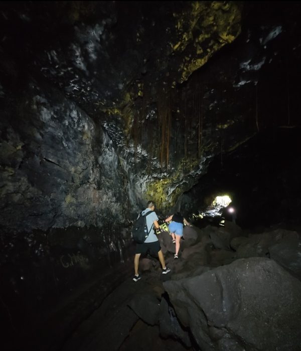 Two explorers navigate rocky terrain inside a dark lava tube cave, with sunlight visible in the distance.