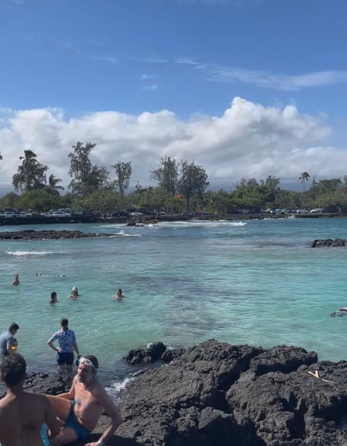 People enjoying a sunny day at a clear blue ocean beach with rocky shores and palm trees.