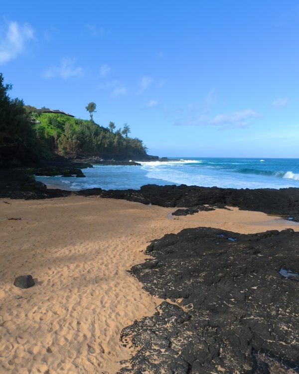 A serene beach scene featuring golden sand, black volcanic rocks, and gentle ocean waves under a clear blue sky.