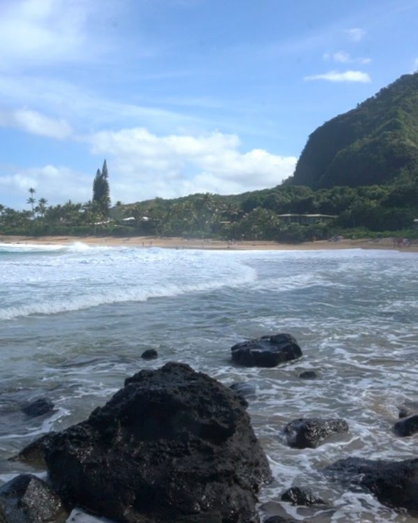 A tranquil beach scene featuring gentle waves lapping against rocky shores, surrounded by lush vegetation and hills under a blue sky.