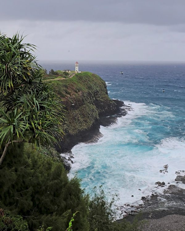 A picturesque view of a lighthouse on a green cliff beside turbulent turquoise waters, framed by lush vegetation under a cloudy sky.