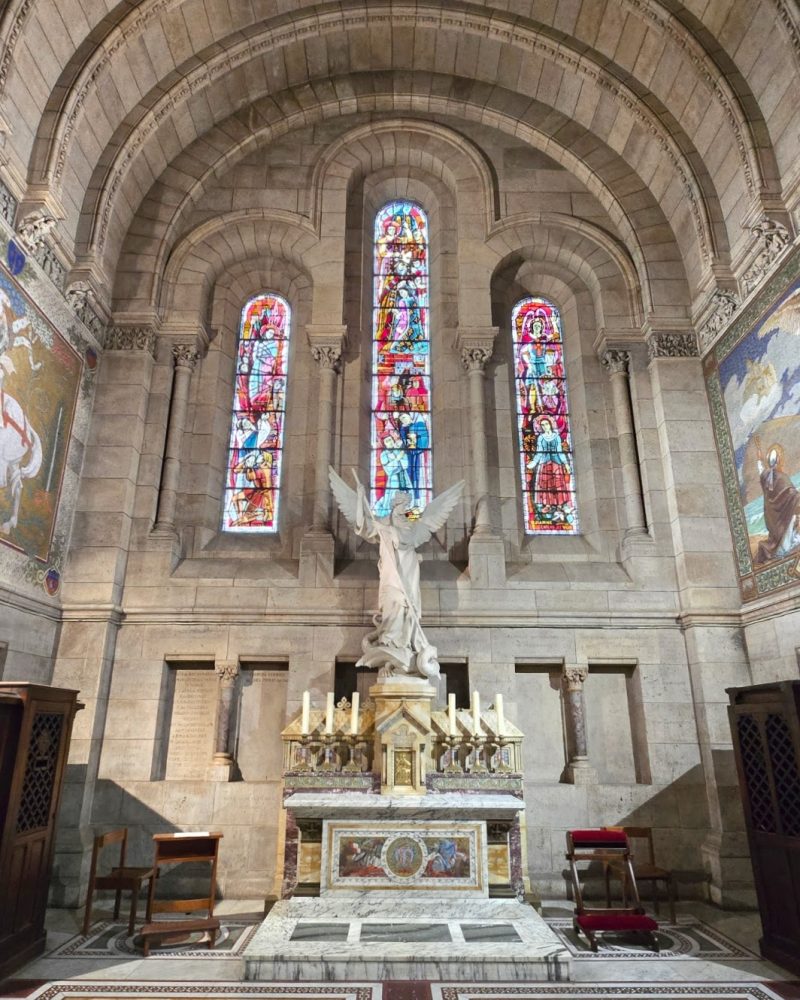 A serene church interior featuring a marble altar with a statue of an angel, surrounded by vibrant stained glass windows and ornate architectural details.