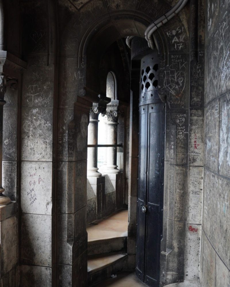 An intricately designed stone corridor featuring a dark door, spiral staircase, and walls etched with graffiti, illuminated by soft natural light.