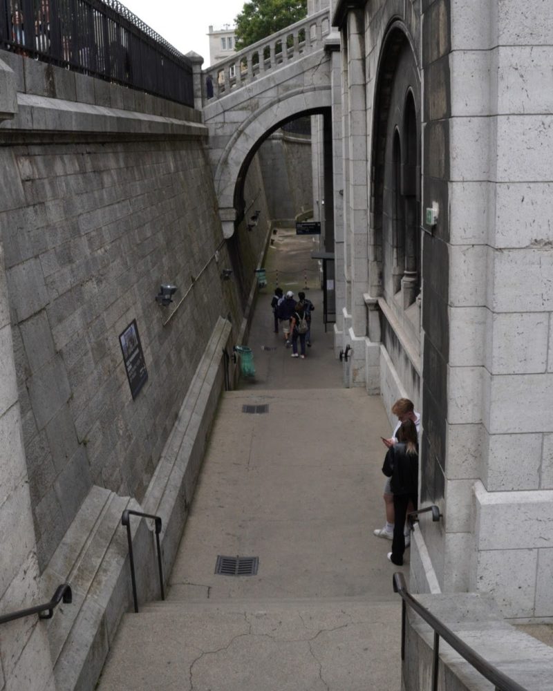 A view of a stairway leading down into a narrow urban walkway, flanked by stone walls and an arched bridge above, with people walking and standing nearby.