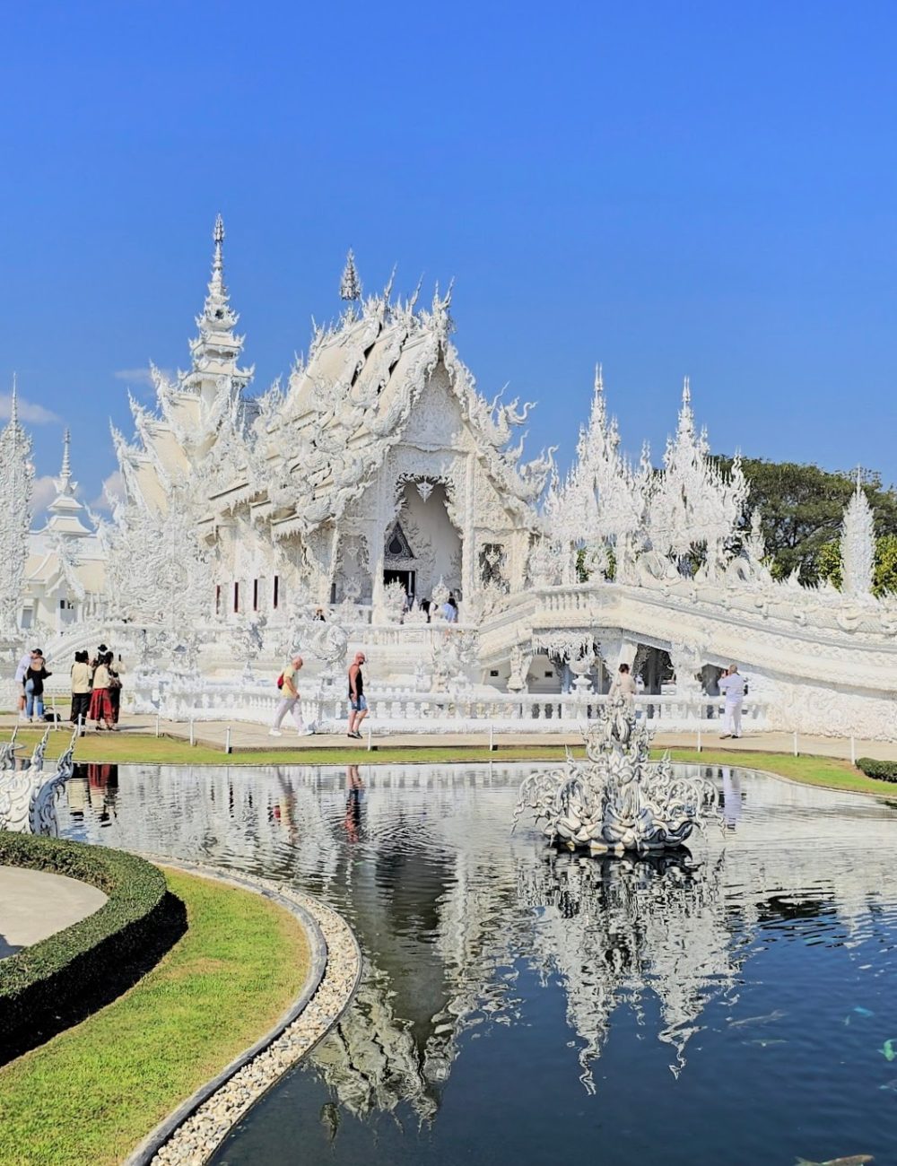 A stunning view of the White Temple in Thailand, showcasing its intricate architecture and reflections in the calm waters, surrounded by lush greenery under a clear blue sky.