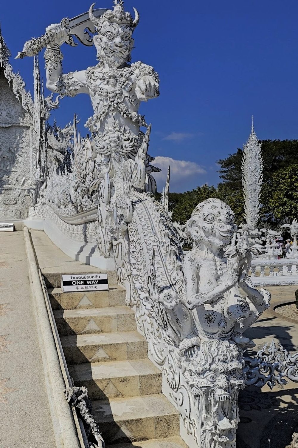 The White Temple, Chiang Rai Intricate white sculptures of mythical figures flank the stairs at Wat Rong Khun, with a sign indicating "One Way" pathway.