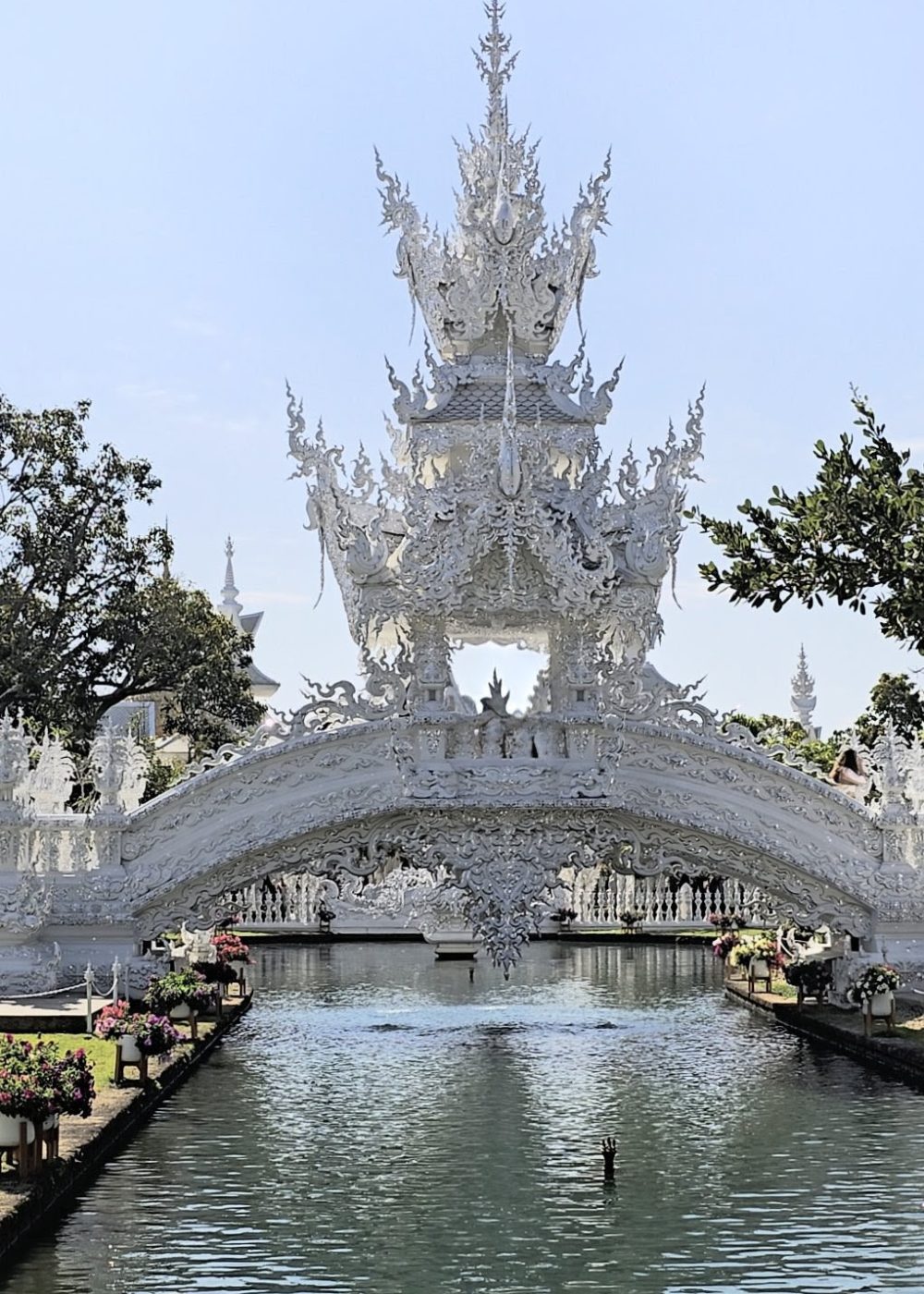A beautifully ornate white bridge adorned with intricate sculptures spanning a serene waterway, surrounded by greenery and bright flowers under a clear sky.