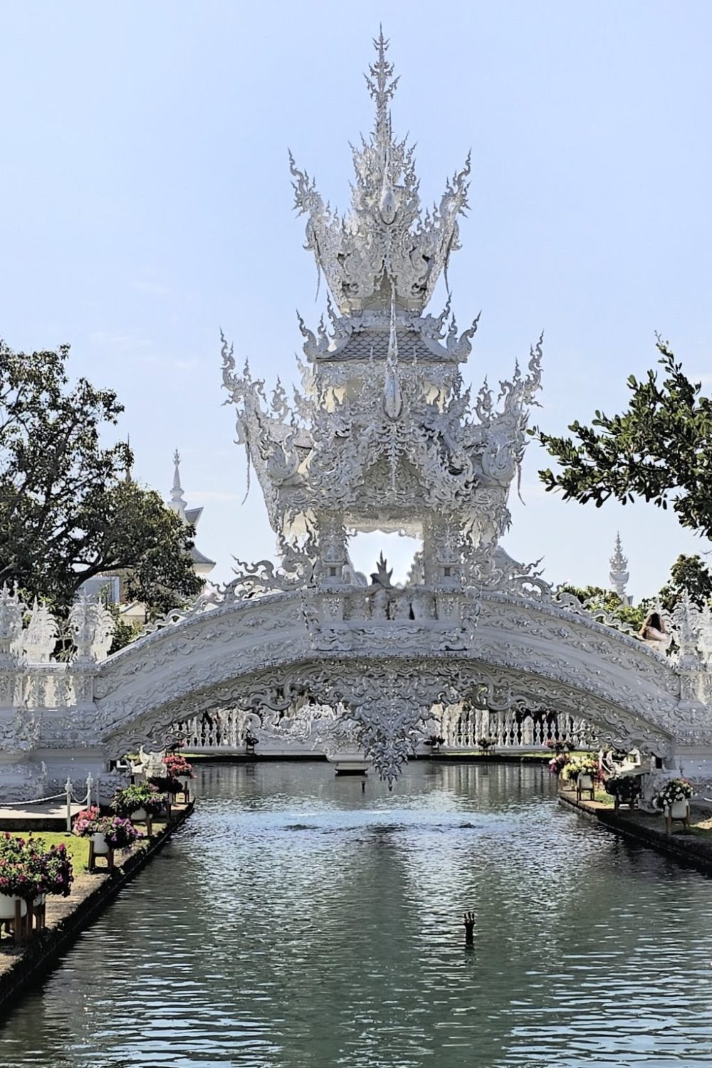 The White Temple, Chiang Rai A beautifully ornate white bridge adorned with intricate sculptures spanning a serene waterway, surrounded by greenery and bright flowers under a clear sky.