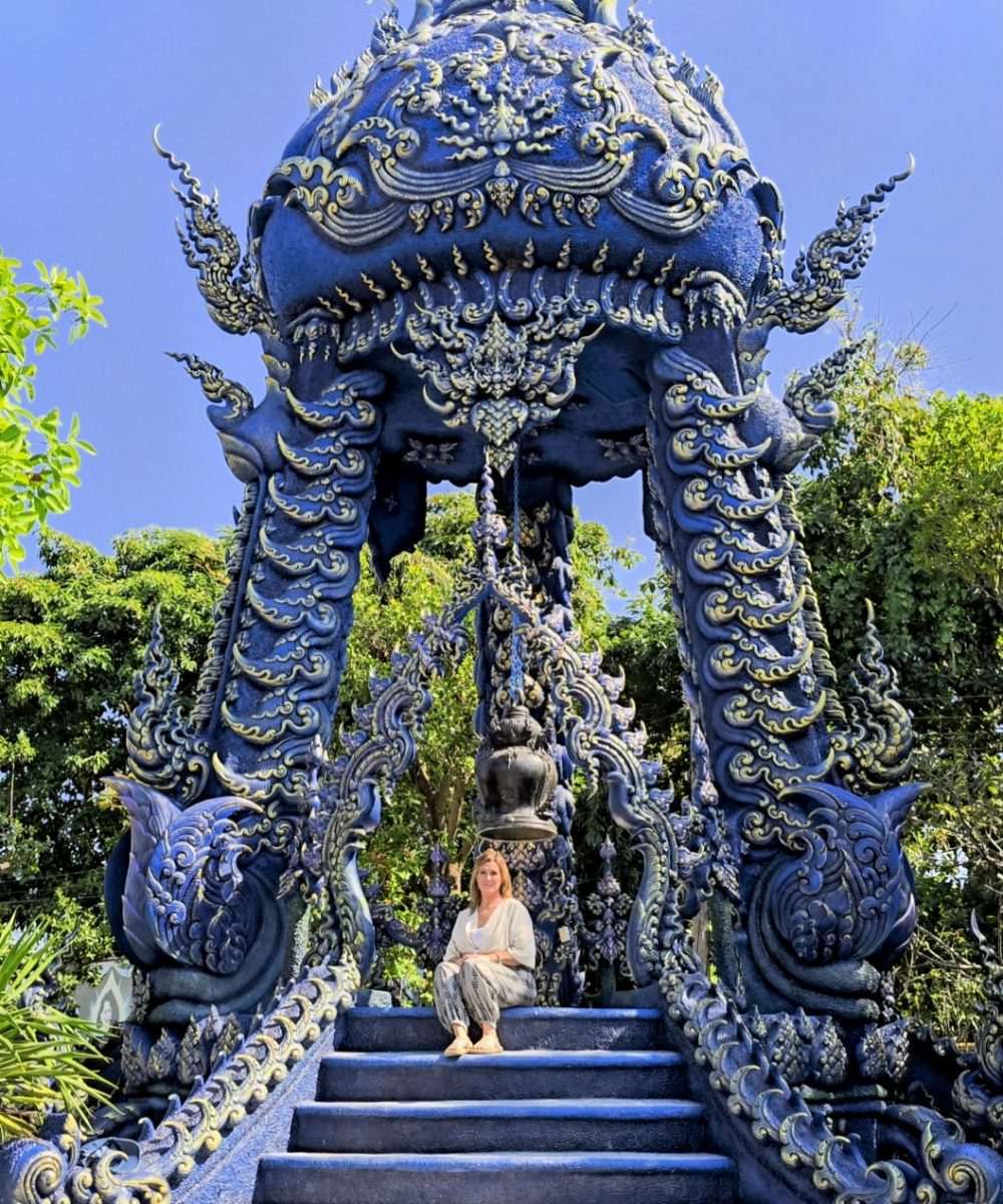 A woman sitting on stairs in front of an intricately designed blue temple surrounded by greenery.