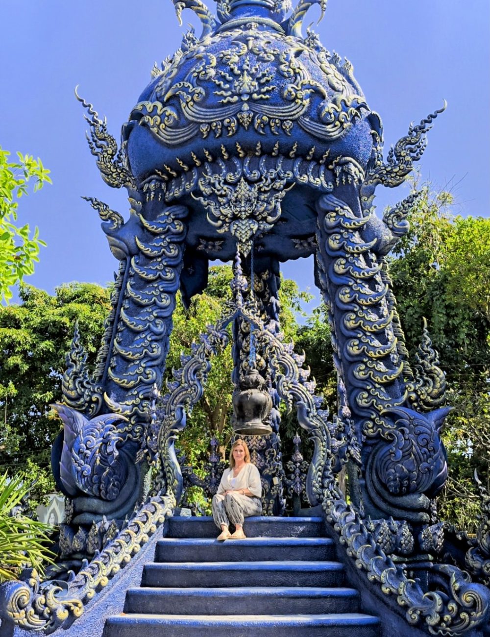 The White Temple, Chiang Rai A woman sitting on stairs in front of an intricately designed blue temple surrounded by greenery.