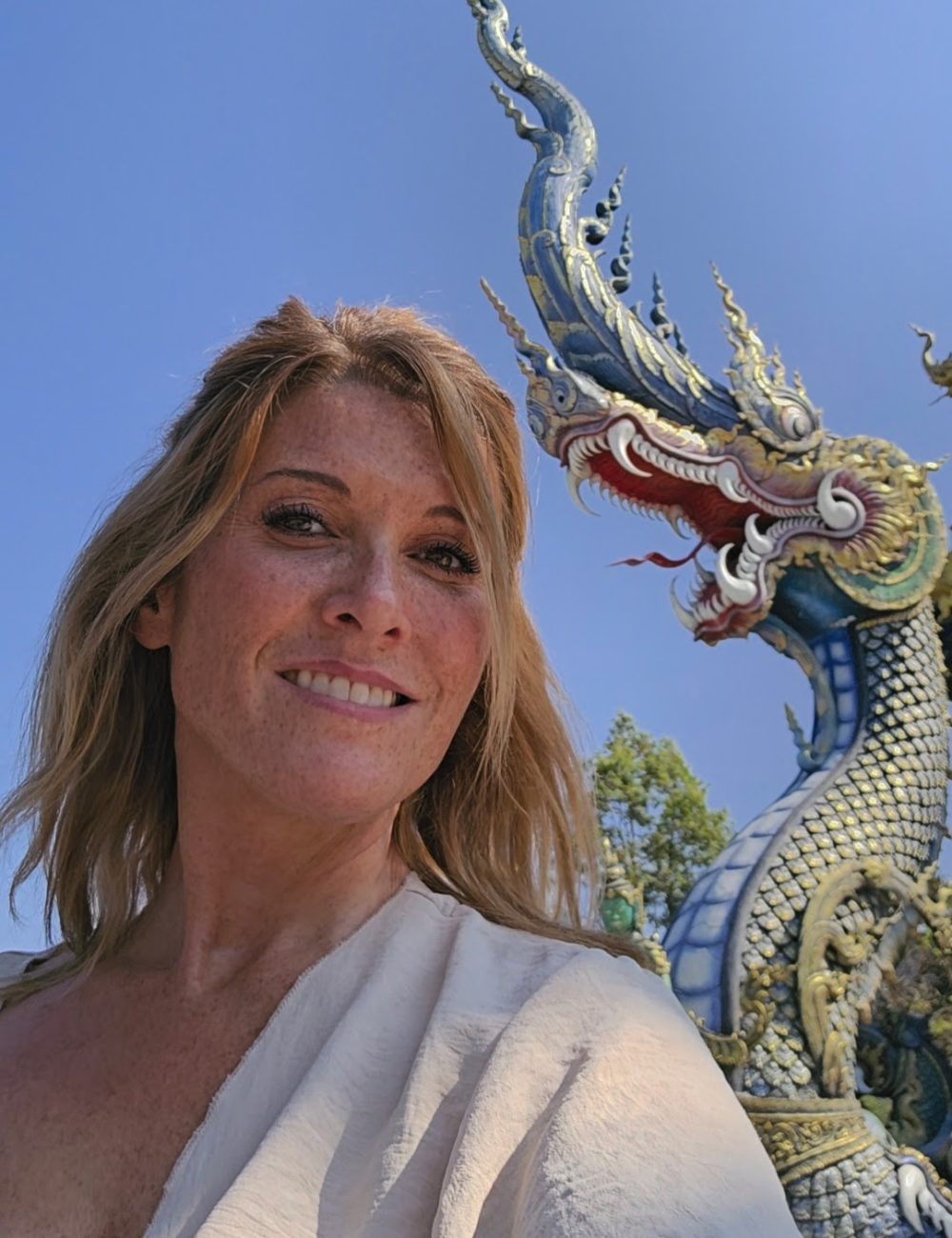 The White Temple, Chiang Rai A woman with long hair smiles in front of a colorful Naga statue against a clear blue sky.