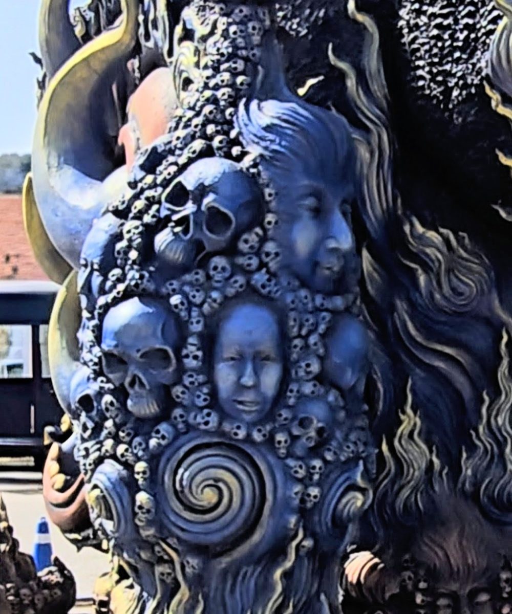 An intricately detailed sculpture featuring skulls and faces intertwined with swirling patterns and vibrant colors, set against a clear blue sky.