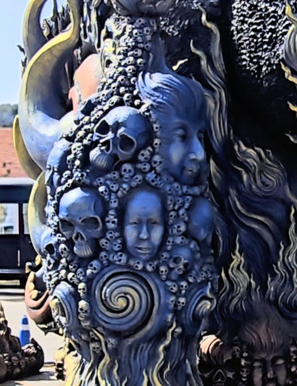 The White Temple, Chiang Rai An intricately detailed sculpture featuring skulls and faces intertwined with swirling patterns and vibrant colors, set against a clear blue sky.