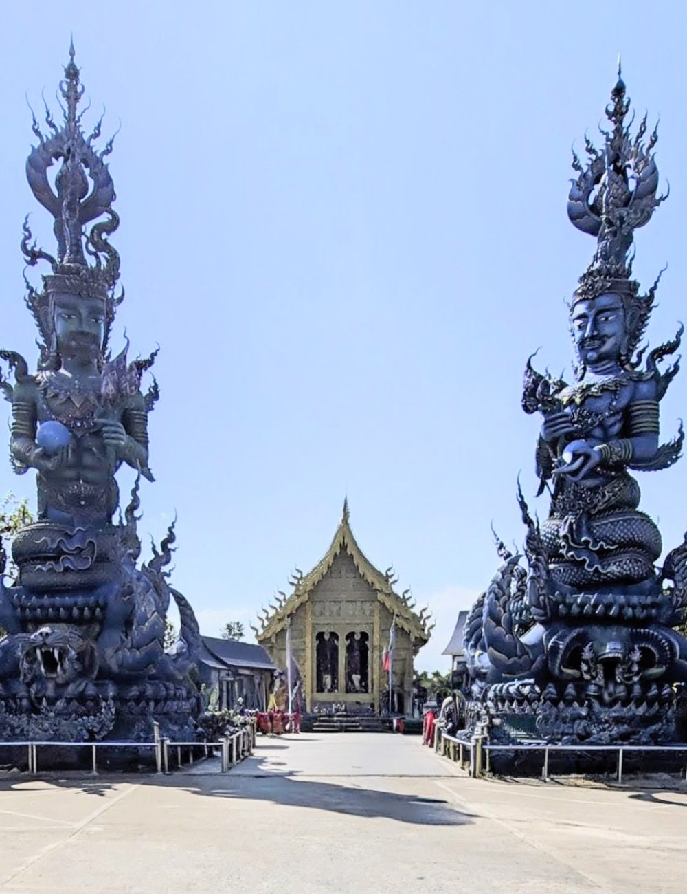 The White Temple, Chiang Rai A stunning view of two large black statues flanking a golden temple, set against a clear blue sky.