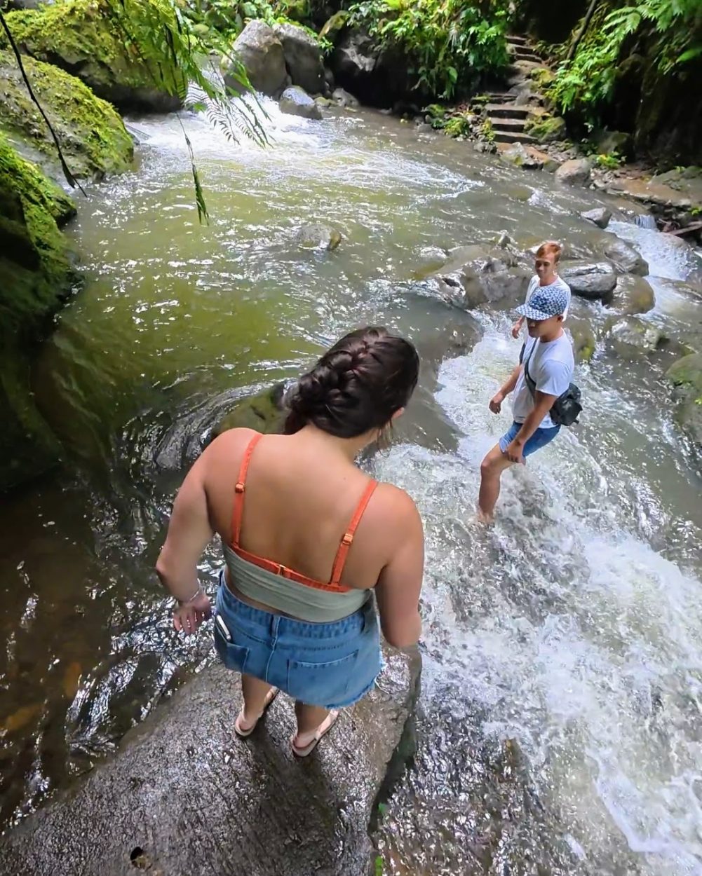 Two friends navigating a rocky stream in a lush, green environment, surrounded by tropical foliage.