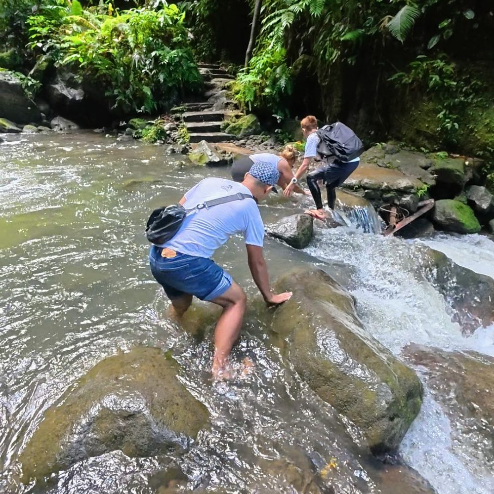 Casaroro Falls — An Adventurous Hike That Was A Little More Than I Expected Three individuals carefully crossing rocky terrain in a lush, green environment near a flowing stream.