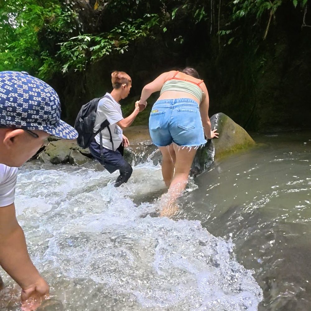 Casaroro Falls — An Adventurous Hike That Was A Little More Than I Expected Two individuals navigate a rocky stream, one helping the other as they wade through the water surrounded by lush greenery.