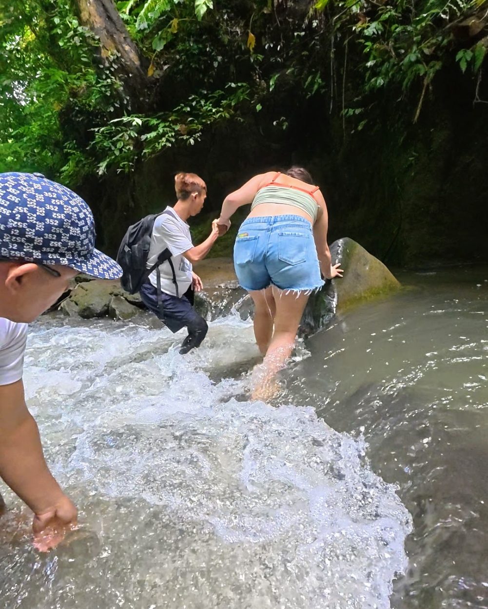 Two individuals navigate a shallow stream with lush green foliage surrounding them, while one helps the other climb over a rock.