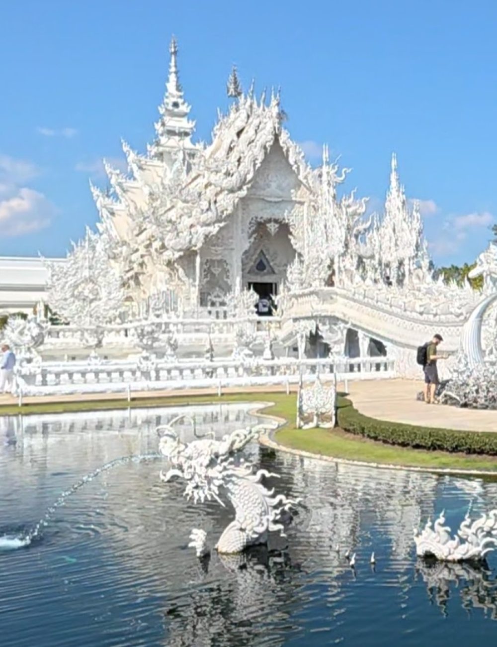 A picturesque view of the White Temple reflecting in a serene pond, surrounded by intricate sculptures and clear blue skies.