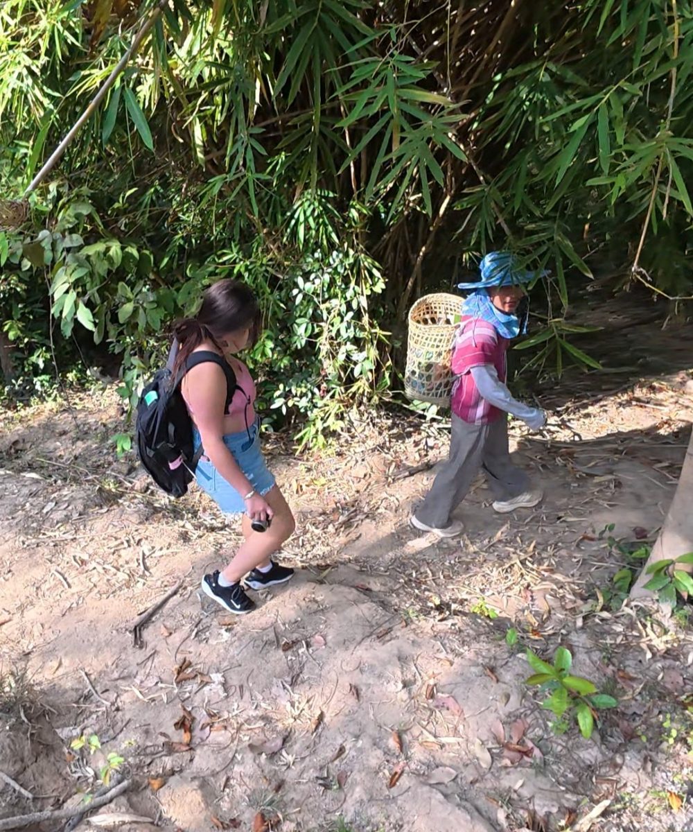 Overnight Stay At Chai Lai Orchid Elephant Sanctuary in Thailand A young woman walks along a dirt path surrounded by lush bamboo greenery, while a man carrying a basket follows her.