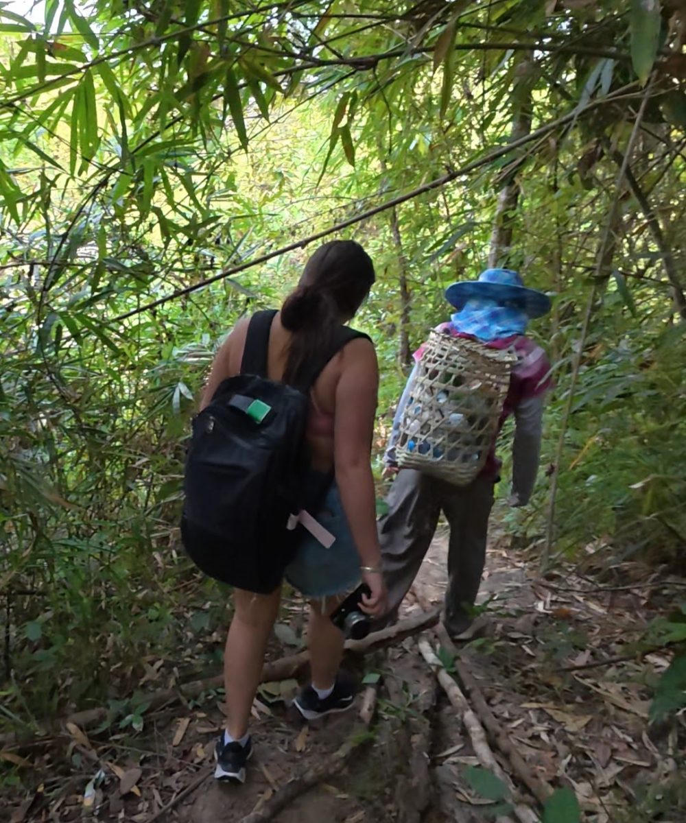 Overnight Stay At Chai Lai Orchid Elephant Sanctuary in Thailand Two individuals navigate a narrow trail surrounded by vibrant bamboo foliage, one carrying a backpack and the other with a woven basket on their back.