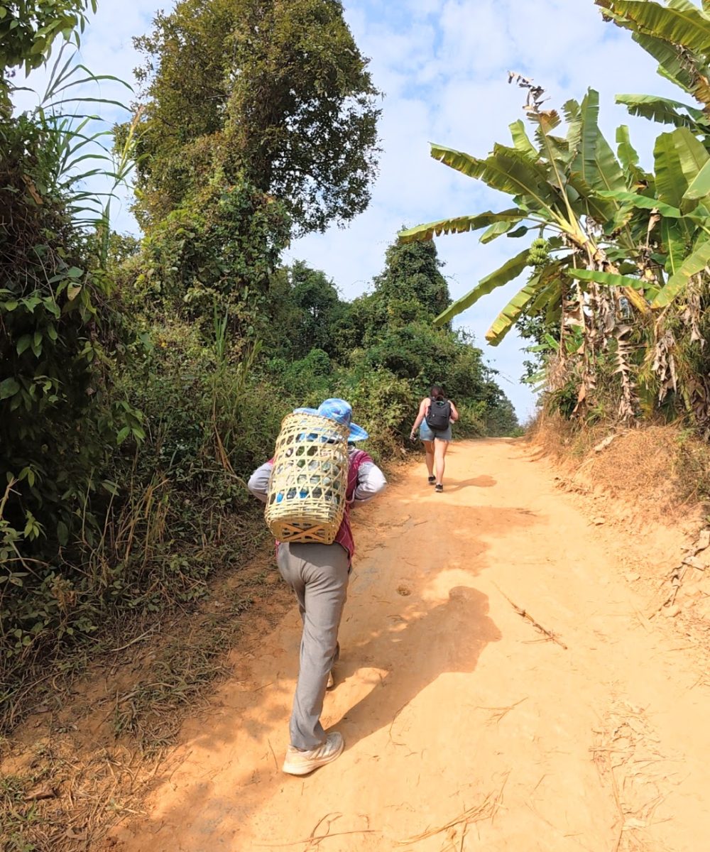 Overnight Stay At Chai Lai Orchid Elephant Sanctuary in Thailand Two hikers ascend a dirt path surrounded by greenery, one carrying a woven basket on their back.