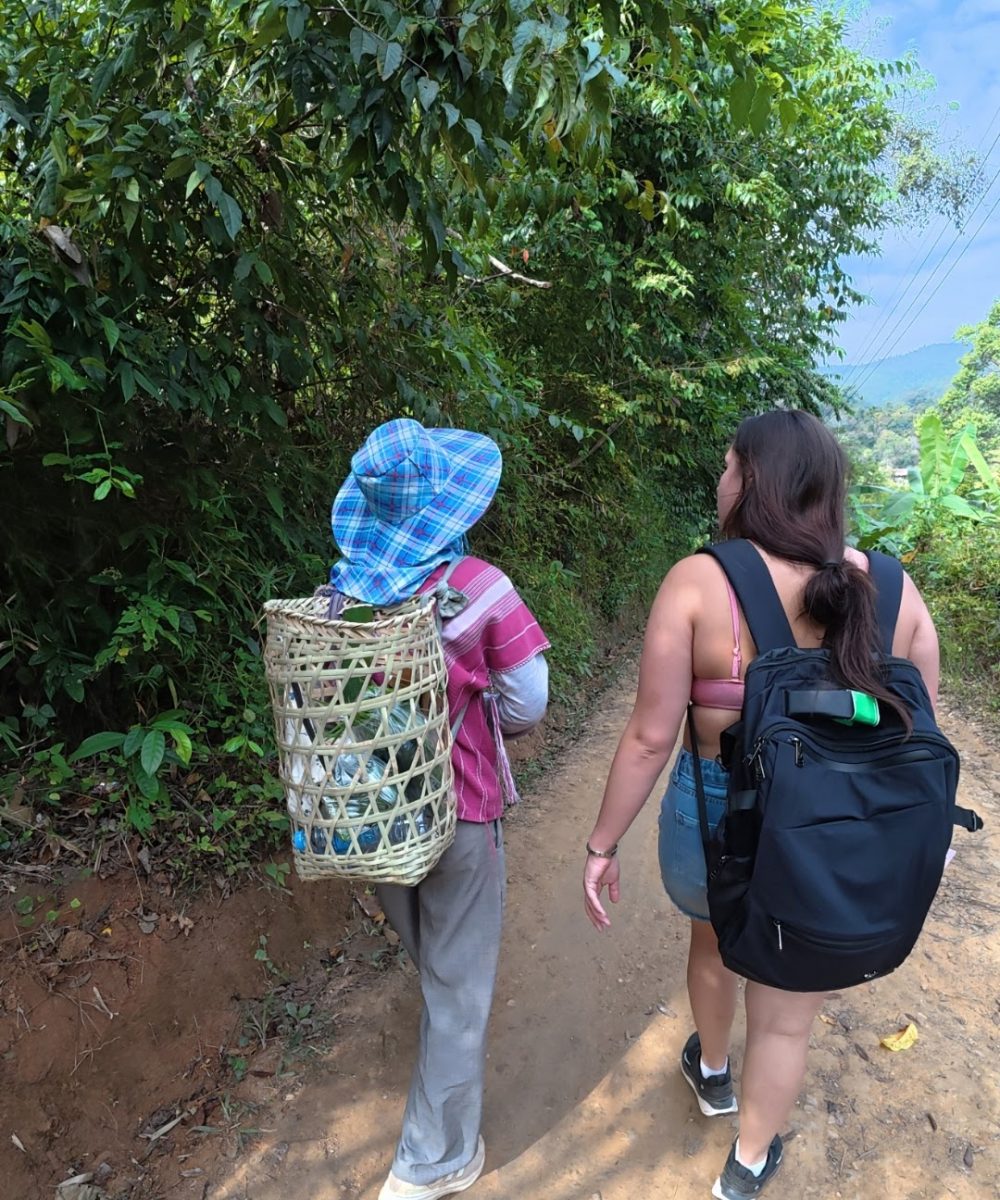 Overnight Stay At Chai Lai Orchid Elephant Sanctuary in Thailand Two women walking along a dirt path surrounded by lush greenery, one wearing a plaid hat and carrying a woven basket, the other with a backpack.