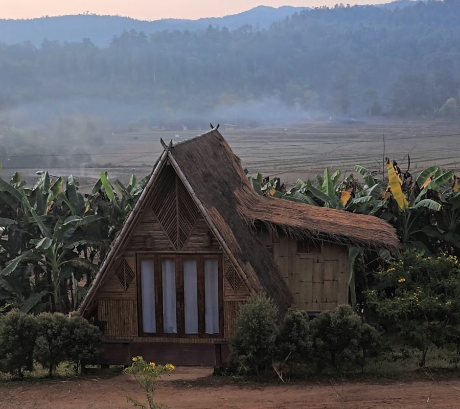 Overnight Stay At Chai Lai Orchid Elephant Sanctuary in Thailand A quaint bamboo cottage surrounded by banana plants, nestled in a tranquil landscape with mountains in the background during dusk.