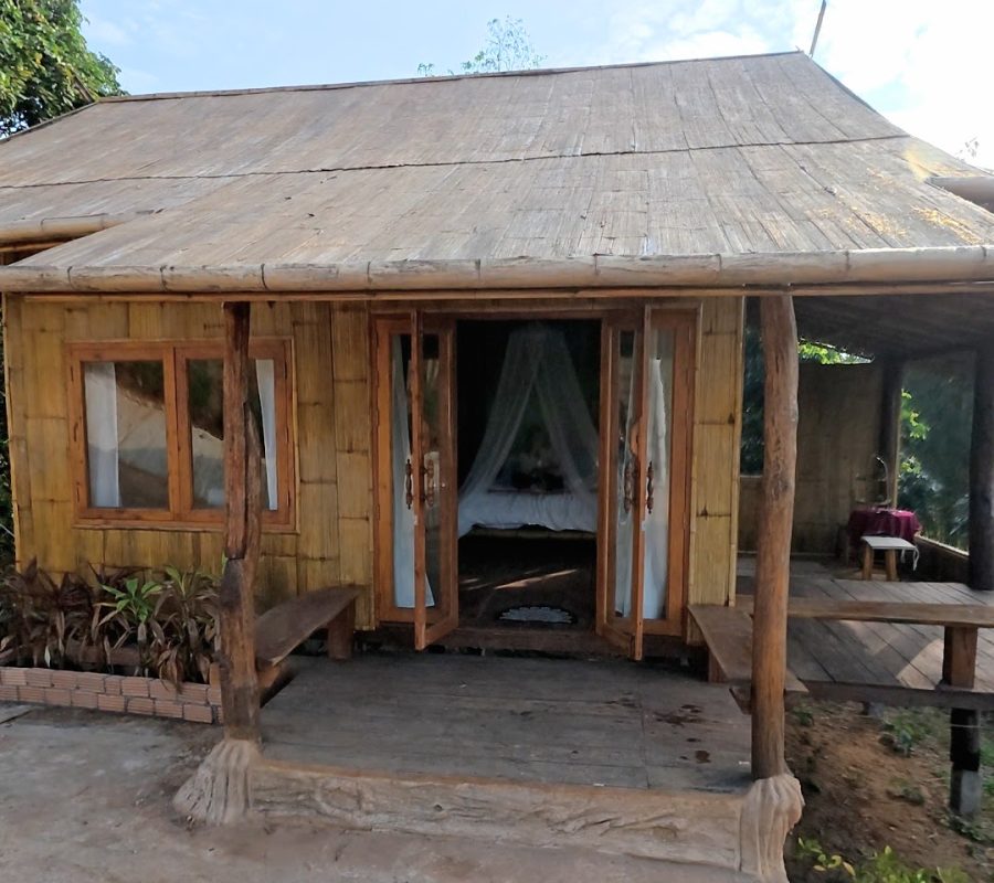 Overnight Stay At Chai Lai Orchid Elephant Sanctuary in Thailand A charming bamboo cottage with open doors, a thatched roof, and a porch surrounded by greenery, inviting a serene outdoor experience.