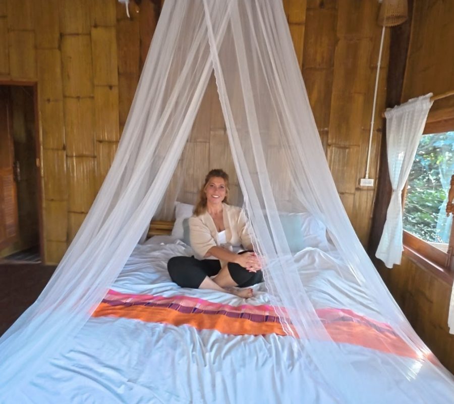 Overnight Stay At Chai Lai Orchid Elephant Sanctuary in Thailand A woman sitting on a bed adorned with a white canopy in a bamboo-themed room, surrounded by natural wood decor and soft lighting.