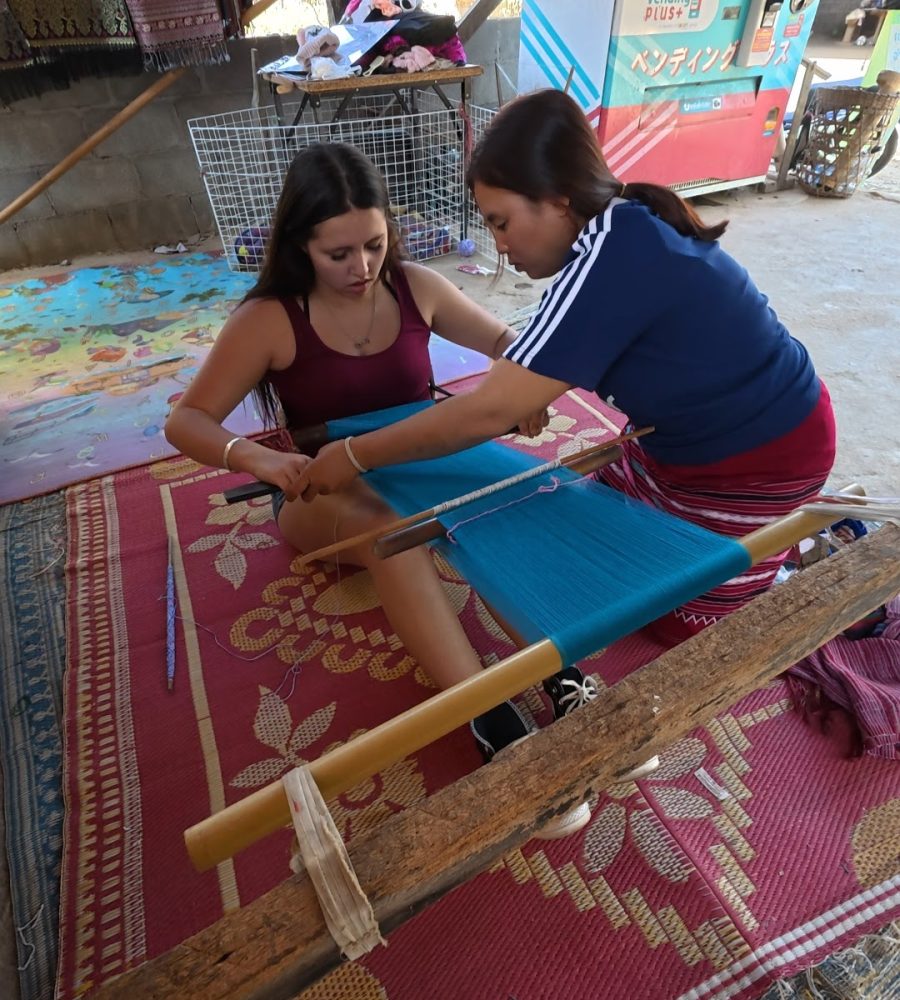Overnight Stay At Chai Lai Orchid Elephant Sanctuary in Thailand Two young women engaged in traditional weaving on a wooden loom, with colorful threads and a vibrant rug beneath them in a sunlit space.