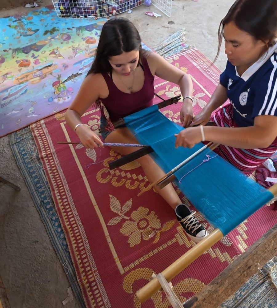 Overnight Stay At Chai Lai Orchid Elephant Sanctuary in Thailand Two women work on a traditional loom, creating a bright blue textile while seated on a colorful patterned rug.