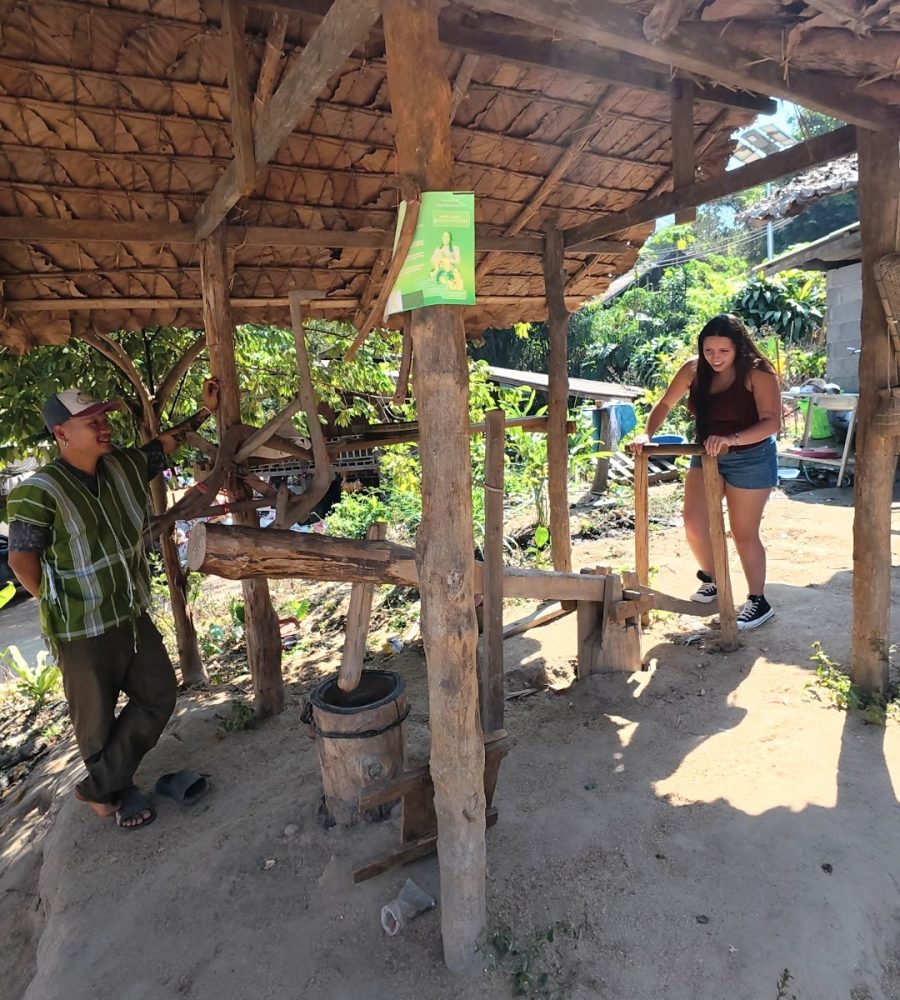 Overnight Stay At Chai Lai Orchid Elephant Sanctuary in Thailand Two individuals interact under a traditional wooden structure, with one operating a hand tool while the other looks on, surrounded by lush greenery.