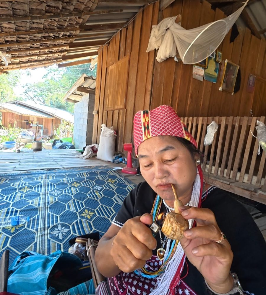 Overnight Stay At Chai Lai Orchid Elephant Sanctuary in Thailand A woman in traditional attire, focused on igniting a decorative pipe while seated indoors, showcasing cultural craftsmanship against a rustic backdrop.