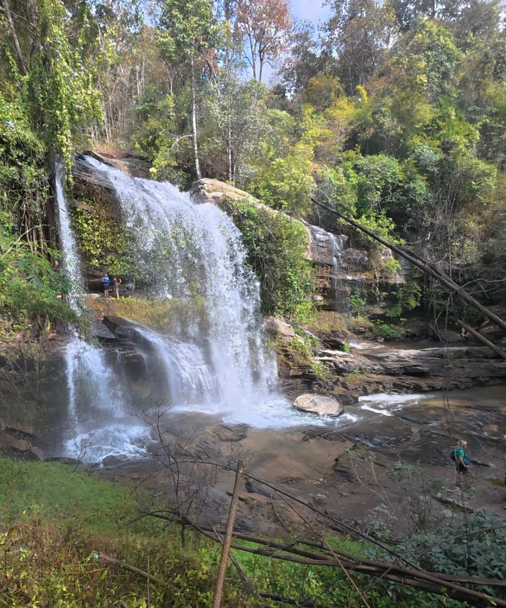 Overnight Stay At Chai Lai Orchid Elephant Sanctuary in Thailand A picturesque waterfall cascading over rocks, with vibrant greenery and two people enjoying the natural scenery nearby.