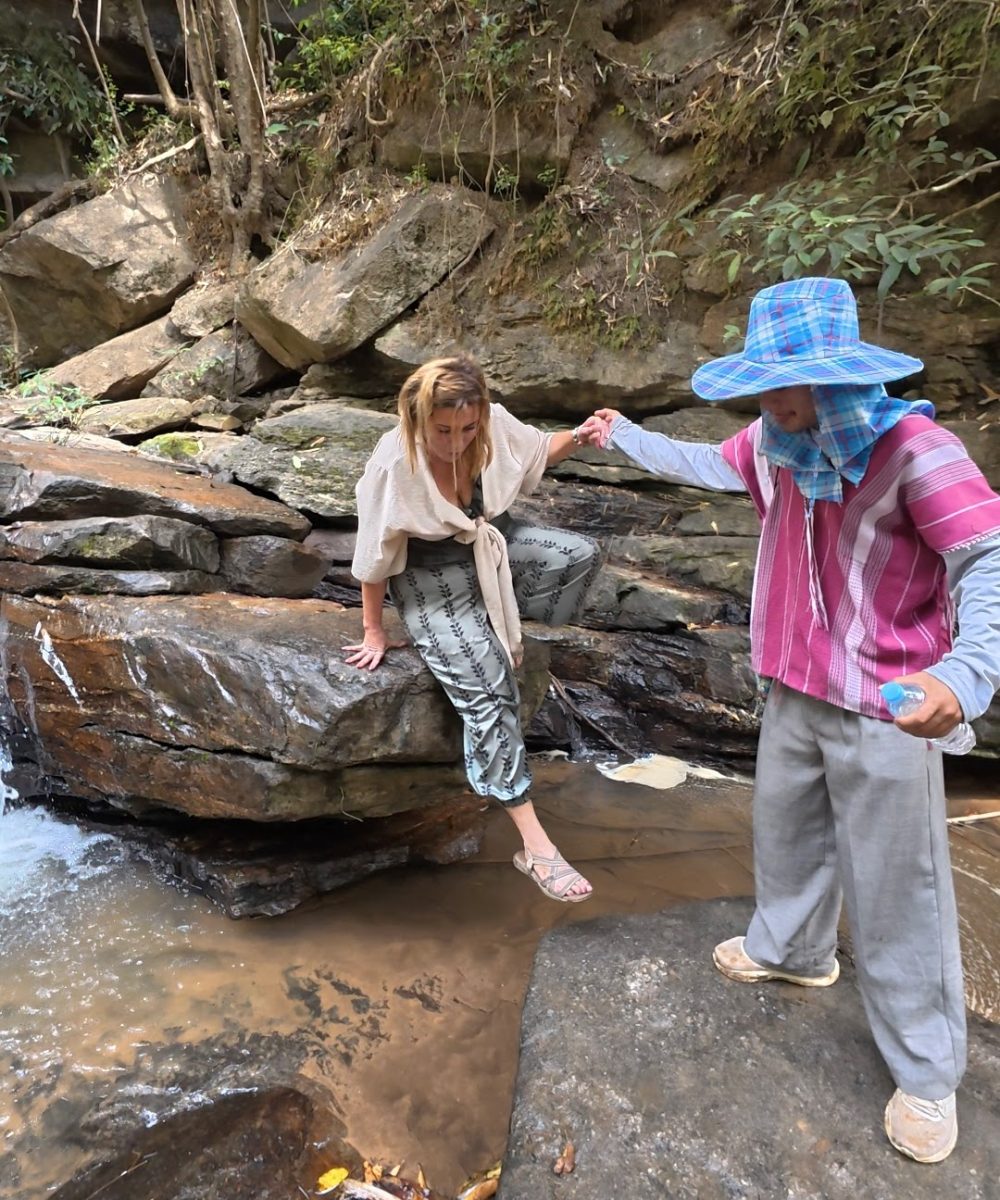 Overnight Stay At Chai Lai Orchid Elephant Sanctuary in Thailand A person helps another navigate rocky terrain near a stream in a natural setting.