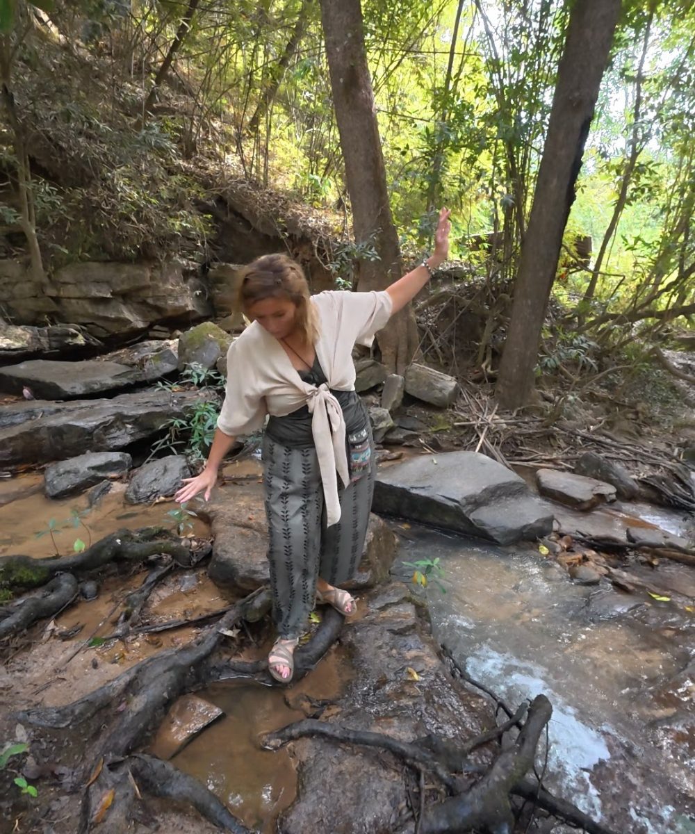 Overnight Stay At Chai Lai Orchid Elephant Sanctuary in Thailand A woman carefully balances on rocks beside a flowing stream in a lush, wooded area.