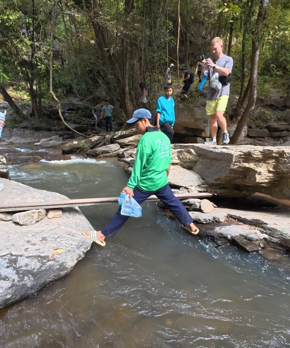Overnight Stay At Chai Lai Orchid Elephant Sanctuary in Thailand A group of people navigate a rocky area near a stream, with one person cautiously crossing a narrow pathway.