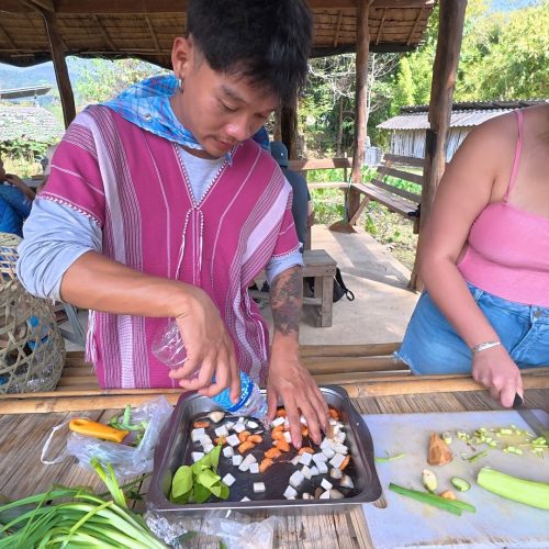 Overnight Stay At Chai Lai Orchid Elephant Sanctuary in Thailand A person in traditional attire prepares fresh ingredients on a tray while another person assists nearby, surrounded by a rustic outdoor setting.