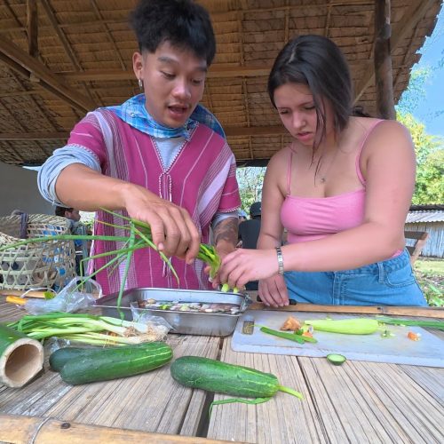 Overnight Stay At Chai Lai Orchid Elephant Sanctuary in Thailand Two individuals collaborating in a rustic kitchen, skillfully preparing vegetables, including cucumbers and green onions, on a bamboo table.