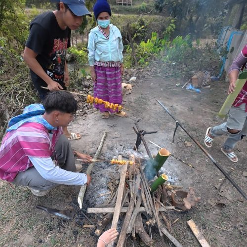 Overnight Stay At Chai Lai Orchid Elephant Sanctuary in Thailand A group of young individuals preparing food over an open fire in a rural setting, with traditional garments and a dog nearby.
