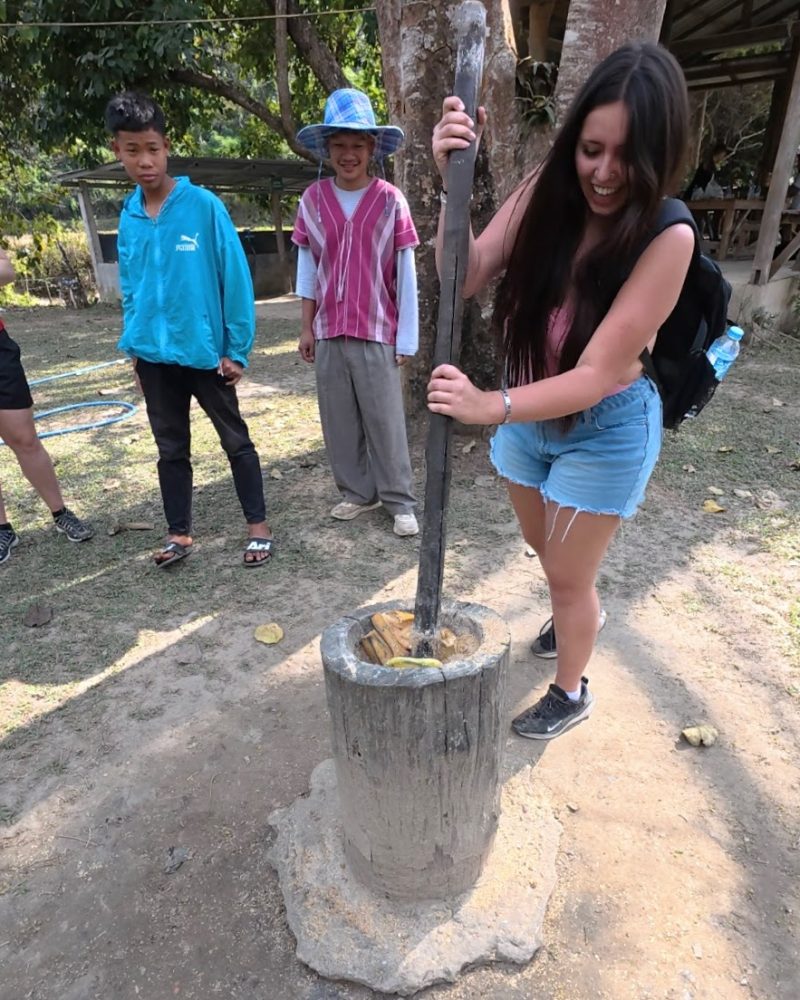 Overnight Stay At Chai Lai Orchid Elephant Sanctuary in Thailand A woman joyfully pounds ingredients in a mortar while others observe, showcasing a traditional cooking method in a rural setting.