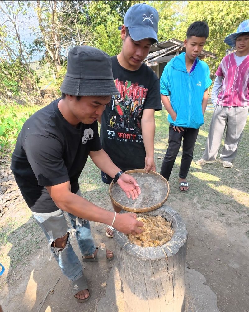 Overnight Stay At Chai Lai Orchid Elephant Sanctuary in Thailand Two young men working together on a traditional food preparation process, surrounded by a group of onlookers in a natural setting.