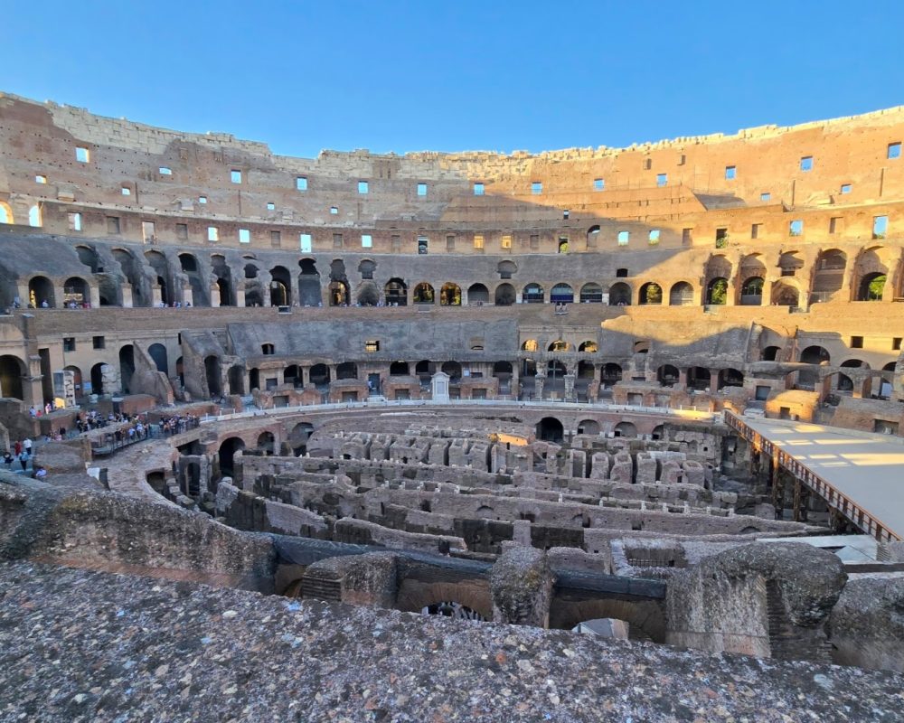 A panoramic view of the interior of the Colosseum showcasing its ancient architecture and seating areas under a clear blue sky.